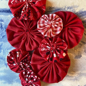 May include: A collection of handmade fabric yo-yos in various sizes and patterns. The yo-yos are primarily red, with some featuring white and black paisley or gingham designs. They are arranged on a blue and white patterned surface.