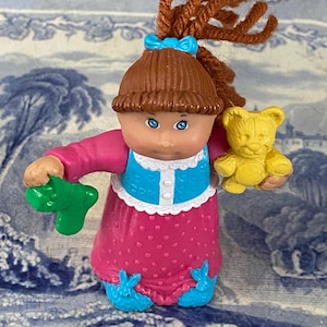 May include: A Cabbage Patch Kid doll wearing a pink dress with white polka dots, a blue and white jacket, and blue bunny slippers. The doll has brown hair, blue eyes, and is holding a green toy and a yellow teddy bear.