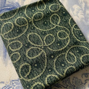 May include: A folded piece of dark forest green fabric featuring a repeating pattern of light cream-colored swirling loops and small dots. The textile has a visible woven texture and is arranged in a rectangular shape with clean, straight edges.