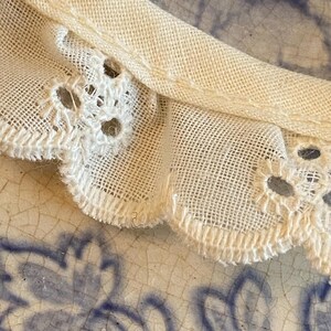 May include: Close-up of a white cotton fabric with a delicate lace trim. The fabric has a slightly textured surface and the lace trim features a scalloped edge.