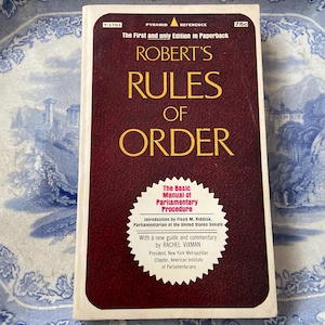 May include: A paperback book titled "ROBERT'S RULES OF ORDER" with a burgundy cover and gold lettering. The book includes the text "The Basic Manual of Parliamentary Procedure" and is set against a blue and white patterned background.