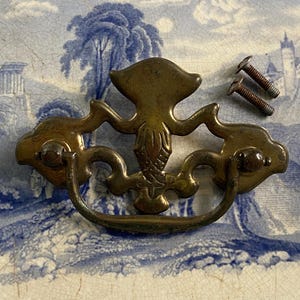May include: Antique brass drawer pull with ornate design, featuring a central decorative element and a curved handle. Includes two screws. The pull is set against a blue and white patterned background.