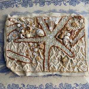 May include: A beige fabric square with a brown embroidered starfish design. The starfish is surrounded by various beads, buttons, and shells.