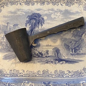 May include: A dark gray metal measuring cup with a long handle, marked with measurements. The handle has the word "Barbour" stamped on it. The cup is set on a blue and white patterned ceramic tray.