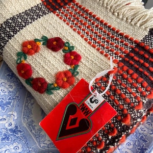May include: A cream-colored woven throw blanket with a red and brown plaid pattern. The blanket has a fringe border and a floral wreath embroidered in red, orange, and green. The blanket has a tag attached that says "Cami-lia" and "MWW".