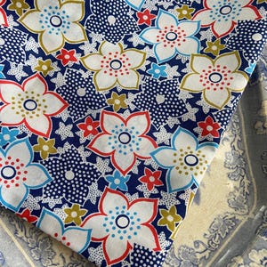 May include: A piece of fabric with a floral pattern. The design features large white flowers with red, blue, and yellow accents, set against a navy blue background with white polka dots. Smaller flowers in red and yellow complete the design.