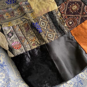 May include: A patchwork textile featuring a variety of fabrics in a range of colors and patterns. The design includes sections of black velvet, satin, and patterned brocade with floral and geometric motifs. The piece rests on a blue and white floral patterned surface.