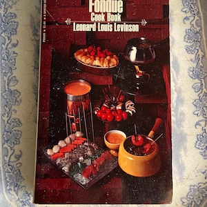 May include: The International Fondue Cook Book by Leonard Louis Levinson. The cover features a colorful illustration of a fondue pot with various dipping items, including fruit, vegetables, and meat.