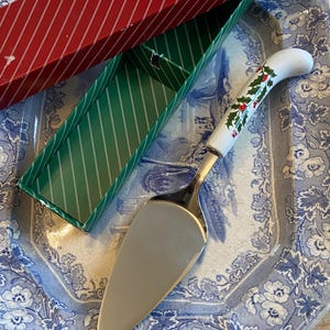 Peut inclure: Une pelle à gâteau argentée avec un manche blanc décoré de houx et de baies rouges. Le serveur est posé sur une assiette à motifs floraux bleus et blancs, avec une boîte à rayures rouges et vertes en arrière-plan. La boîte porte le mot "Cummings".