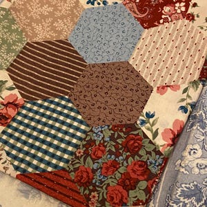 May include: A patchwork quilt featuring a variety of hexagonal fabric pieces in diverse patterns and colors, including red floral, blue vine, brown polka dot, striped, and green gingham prints, all stitched together on a light fabric base.
