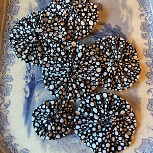 May include: Six black and white fabric coasters with a polka dot design. The round coasters have a gathered centre, resembling small flowers. They are arranged on a decorative blue and white plate.