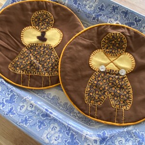 May include: Two brown, round fabric pot holders with yellow and floral accents. One features a dress design, the other a shorts design, both with button details. The pot holders are on a blue and white floral patterned tray.