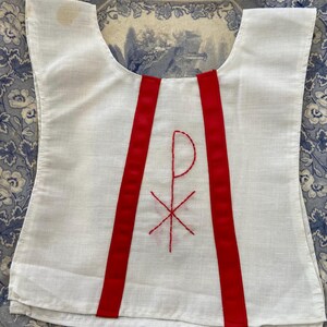 May include: White linen vest with two red ribbon straps and a red embroidered symbol of a cross and a letter P.
