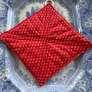 May include: A red, diamond-shaped quilted pot holder with a white floral pattern and a white hanging loop. It rests on a blue and white floral patterned plate.