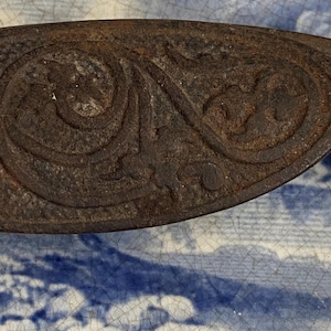 Puede incluir: Un tope de puerta de hierro fundido vintage con un diseño floral decorativo. El tope de puerta es marrón y tiene una superficie lisa y desgastada.