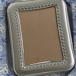 May include: A silver picture frame with a heart-shaped border. The frame is sitting on a blue and white patterned surface.