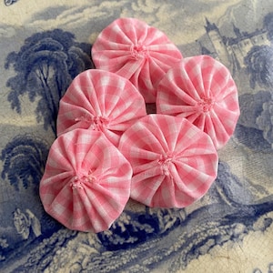 May include: Six pink and white gingham fabric yo-yos, each with a gathered centre, resembling small flowers. They are arranged on a blue and white patterned surface, creating a vintage aesthetic. The yo-yos are a charming craft item.