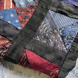 May include: A patchwork quilt made from various patterned neckties in shades of blue, red, and black. The quilt is constructed with triangular pieces and bordered with black ribbon. The quilt is placed on a blue and white floral patterned surface.