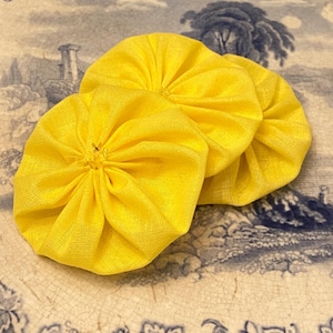 May include: Three vibrant yellow fabric flowers, each with a gathered, circular form. The flowers are made from a textured material and are placed on a vintage plate with a blue and white floral design.
