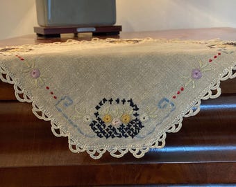 May include: A beige linen doily with a scalloped edge and embroidered floral designs in shades of blue, yellow, red, and black.