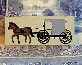May include: A rectangular wooden block features a painted illustration of a horse-drawn carriage. The horse is brown with a dark mane and tail, pulling a gray carriage with black wheels. The background is a cream color.