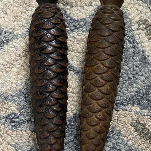 May include: Two antique, dark brown metal pine cone ornaments. Each has a loop at the top for hanging and a textured, scaled surface. The pine cones are displayed on a patterned, neutral-toned surface.