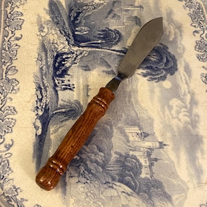 May include: A butter knife with a wooden handle and a stainless steel blade rests on a blue and white patterned plate. The plate has a landscape design. The knife's handle is a warm brown color.