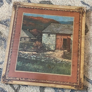 May include: Framed landscape print featuring a rustic barn with a red door and a stone exterior. The artwork depicts a rural scene with a log cabin, a stone wall, and a mountain backdrop. The frame has a decorative, ornate design.