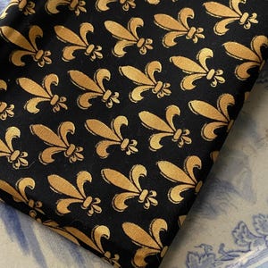 May include: Folded black fabric featuring a repeating gold fleur-de-lis pattern. The fabric is a dark colour with a repeating pattern of a stylized lily flower design. The fabric is folded, showing the pattern at an angle.