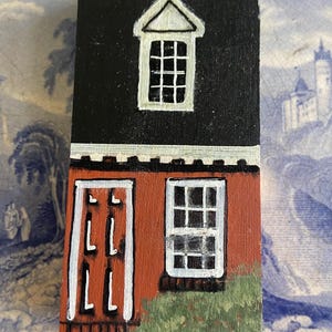 May include: A hand-painted wooden block depicting a two-story house. The top story has a black roof and a white-framed window. The bottom story is red with a white-framed window and door. Green bushes are painted at the base.
