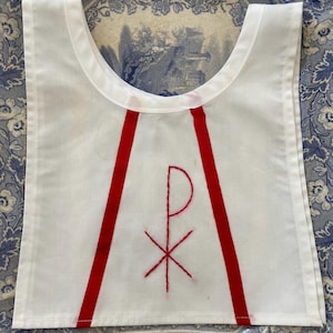 May include: White bib with red accents and embroidered symbol. The bib features two red vertical stripes and a red embroidered symbol in the center. The bib is made of white fabric.