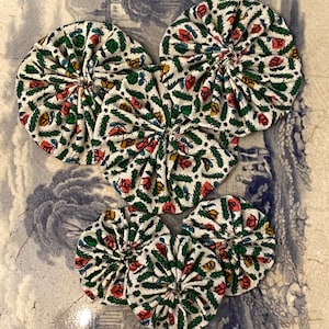 May include: Six fabric yo-yos with a floral design. The yo-yos are white with a repeating pattern of red, yellow, and green flowers and leaves. They are arranged on a blue and white patterned surface.