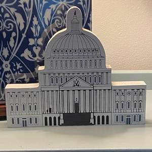 Op de afbeelding: Een wit houten model van een gebouw met een koepelvormig dak en kolommen. Het gebouw heeft een grijs en zwart ontwerp met ramen en een deur.