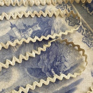 May include: Close-up of cream-coloured, zig-zag ribbon. The ribbon is placed on a vintage blue and white patterned plate. The ribbon's texture appears soft and slightly textured, suitable for craft projects or adding a decorative touch.