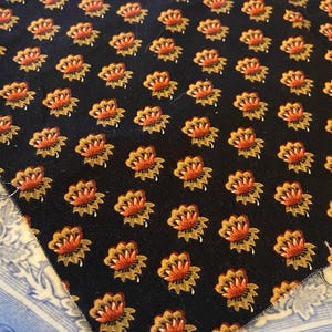May include: A piece of black fabric featuring a repeating pattern of small, stylized flowers with orange petals and gold-toned leaves. The textile has a consistent, evenly spaced floral print across the entire visible surface area.