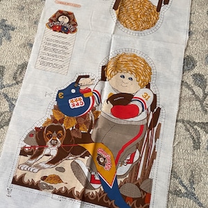 May include: A white fabric panel featuring a printed Cabbage Patch Kids sewing pattern. The design shows a child with blonde hair wearing a football uniform, holding a football and a blue helmet, with a dog and a pennant. Includes instructions.