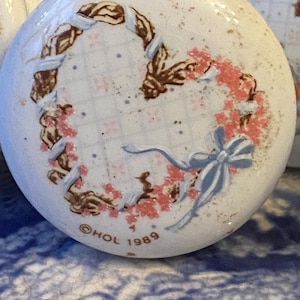 May include: White ceramic knob with a heart-shaped design. The heart is made of a brown rope-like material with pink flowers and a blue ribbon. The knob is marked with the text "HOL 1989".