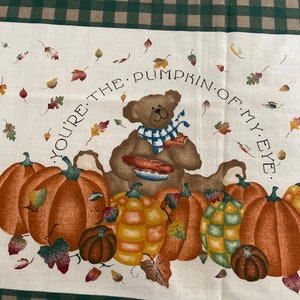 Peut inclure: Un panneau de tissu blanc avec un motif à carreaux vert et marron. Le panneau présente un ours en peluche brun tenant une tarte, entouré de citrouilles et de feuilles d'automne. Le texte "You're the pumpkin of my eye." est écrit en cercle autour de l'ours.