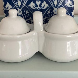 May include: A white ceramic salt and pepper shaker set with two round compartments and matching domed lids. The set has a handle and sits on a light blue surface. A blue and white patterned object is in the background.