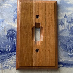 May include: A rectangular, wooden light switch cover with a natural wood grain finish. The cover has a rectangular opening for the switch and two screw holes. The wood is a warm, medium brown color.