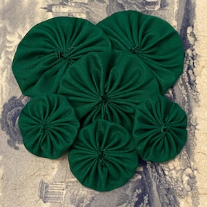 May include: Six dark green fabric yo-yos of different sizes are displayed on a patterned surface. The yo-yos have a gathered, circular design with a central point. The fabric is a solid colour, and the yo-yos are evenly spaced across the surface.