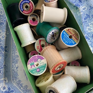 May include: A green box filled with vintage spools of thread in various colors, including white, green, black, and tan. The spools have labels with text like "Coats & Clarks" and "Mercerized Cotton". The background is a blue and white floral pattern.