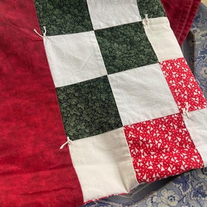 May include: A patchwork quilt with a red border and a checkerboard pattern of white, green floral, and red triangle print squares. The quilt is resting on a blue floral patterned plate.