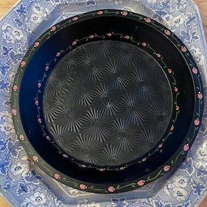 May include: A black pie pan with a floral design around the rim, sitting on a blue and white patterned plate. The pan has a textured interior and the text "OVENEX 11-310-10 USA". The floral design features pink flowers and green vines.