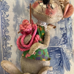 May include: A handmade fabric mouse doll with a floral painted face, wearing a green floral dress and carrying a pink rope bundle. The doll has stitched details and a black pom-pom nose. The ears are pink.