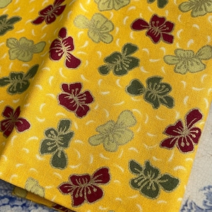 May include: A folded yellow fabric with a repeating floral pattern. The flowers are in shades of burgundy and olive green, with a subtle white swirl design. The fabric has a textured appearance.