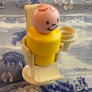 May include: A yellow and white plastic toy figure of a person sitting in a chair with a cup in their hand. The figure has a pink head with red hair and a black smiling face.