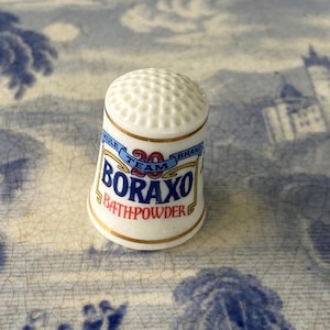 Può includere: Un ditale in porcellana bianca con la parte superiore a pois e finiture dorate. Il ditale presenta il testo "BORAXO BATHPOWDER" in blu e rosso, con "MULE TEAM BRAND" in un design a banner.