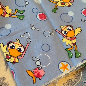 May include: Light blue fabric with a repeating pattern of cartoon ducks wearing snorkels and flippers, along with fish and bubbles. The ducks are yellow with orange beaks and red accents. The fish are orange and pink. The bubbles are white.