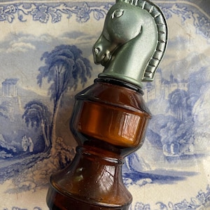May include: A vintage brown glass bottle with a silver horse head stopper. The bottle has a rounded base and a wide neck. The horse head is detailed with a mane and a bridle.